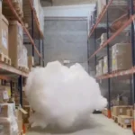 Um potente gerador de nevoeiro Density HP14000 IP fixado a um pilar industrial, projetando uma imensa e densa nuvem de fumo branco que enche instantaneamente todo o volume e a altura do teto de um grande armazém logístico, sob o olhar atento de um técnico.
