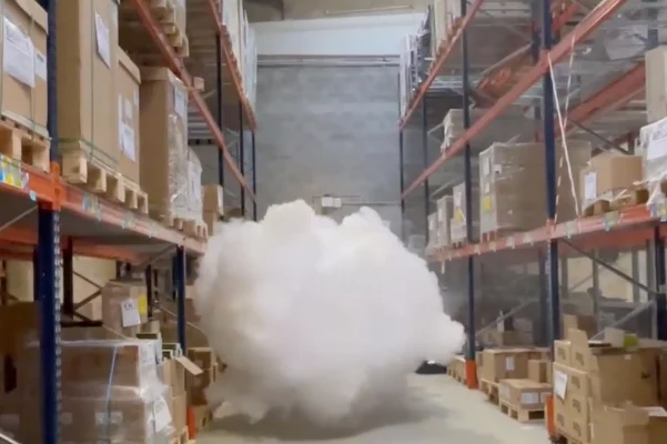 Un puissant générateur de brouillard Density HP14000 IP fixé sur un pilier industriel, projetant un immense et dense nuage de fumée blanche qui sature instantanément tout le volume et la hauteur sous plafond d'un grand entrepôt logistique, sous le regard d'un technicien.