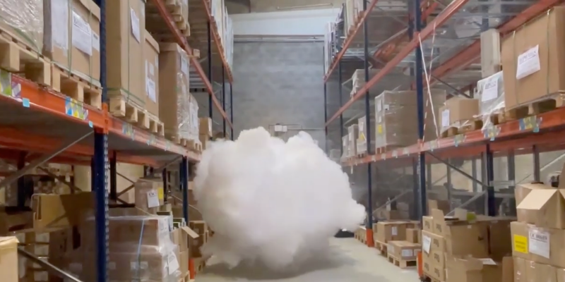 Un puissant générateur de brouillard Density HP14000 IP fixé sur un pilier industriel, projetant un immense et dense nuage de fumée blanche qui sature instantanément tout le volume et la hauteur sous plafond d'un grand entrepôt logistique, sous le regard d'un technicien.